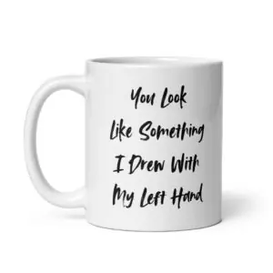 Funny sarcastic white ceramic coffee mug with black brush script typography reading "YOU LOOK LIKE SOMETHING I DREW WITH MY LEFT HAND" centered on the side.