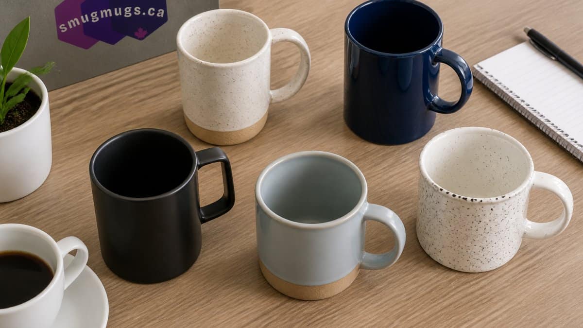 Overhead shot of five ceramic mugs in different colors and finishes arranged on a light wooden table, each displaying distinct branding styles, with a coffee cup and notepad nearby